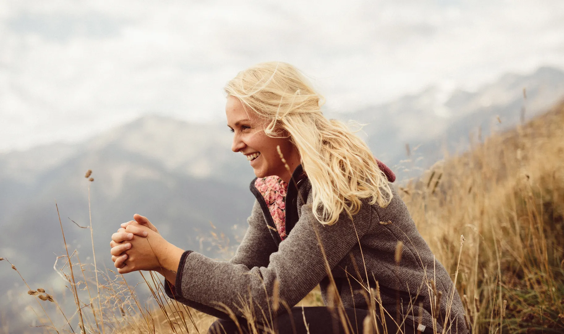 Blonde Frau in grauer Strickjacke aus Schurwolle lächelt seitlich in herbstlicher Berglandschaft