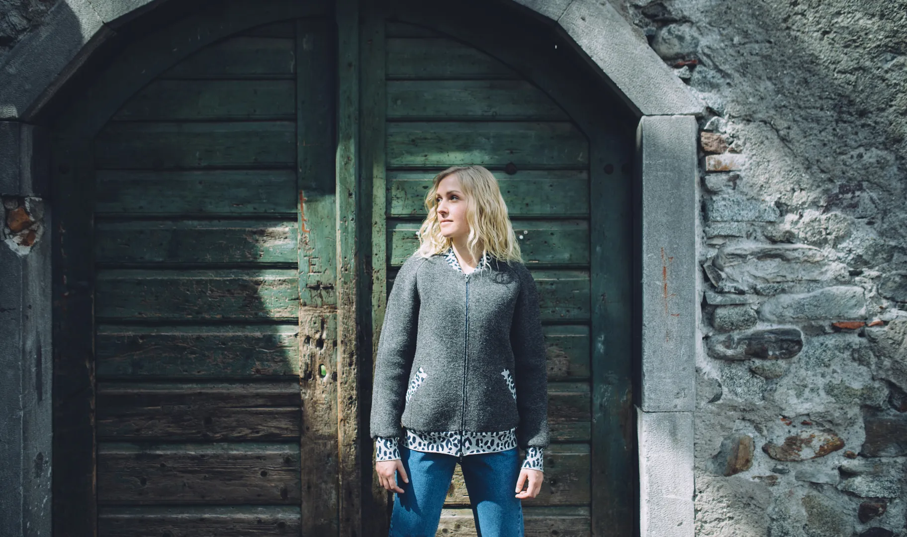 Blonde Frau in grauem Strickjanker mit Reißverschluss vor altem Holztor und Steinmauer