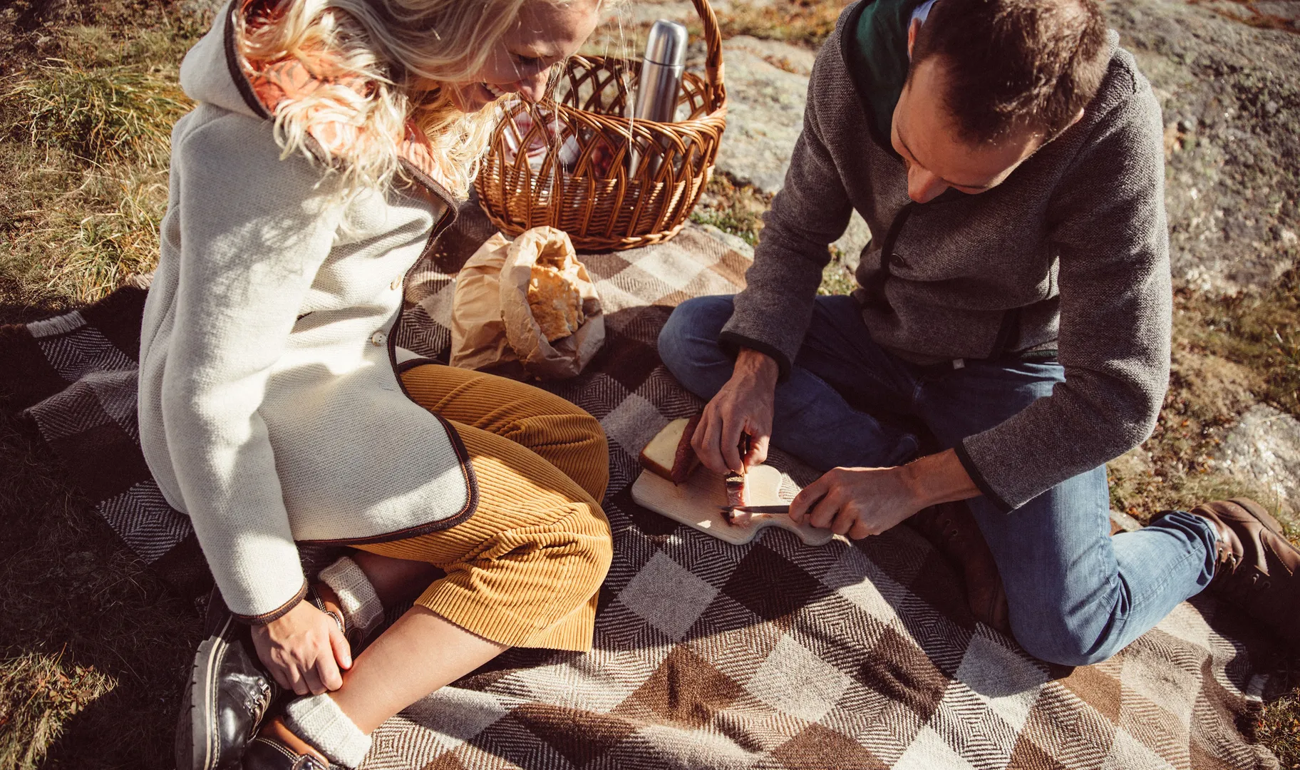 Paar bei Picknick auf karierter Wolldecke mit Walkjacken, Brot und Korb in den Bergen
