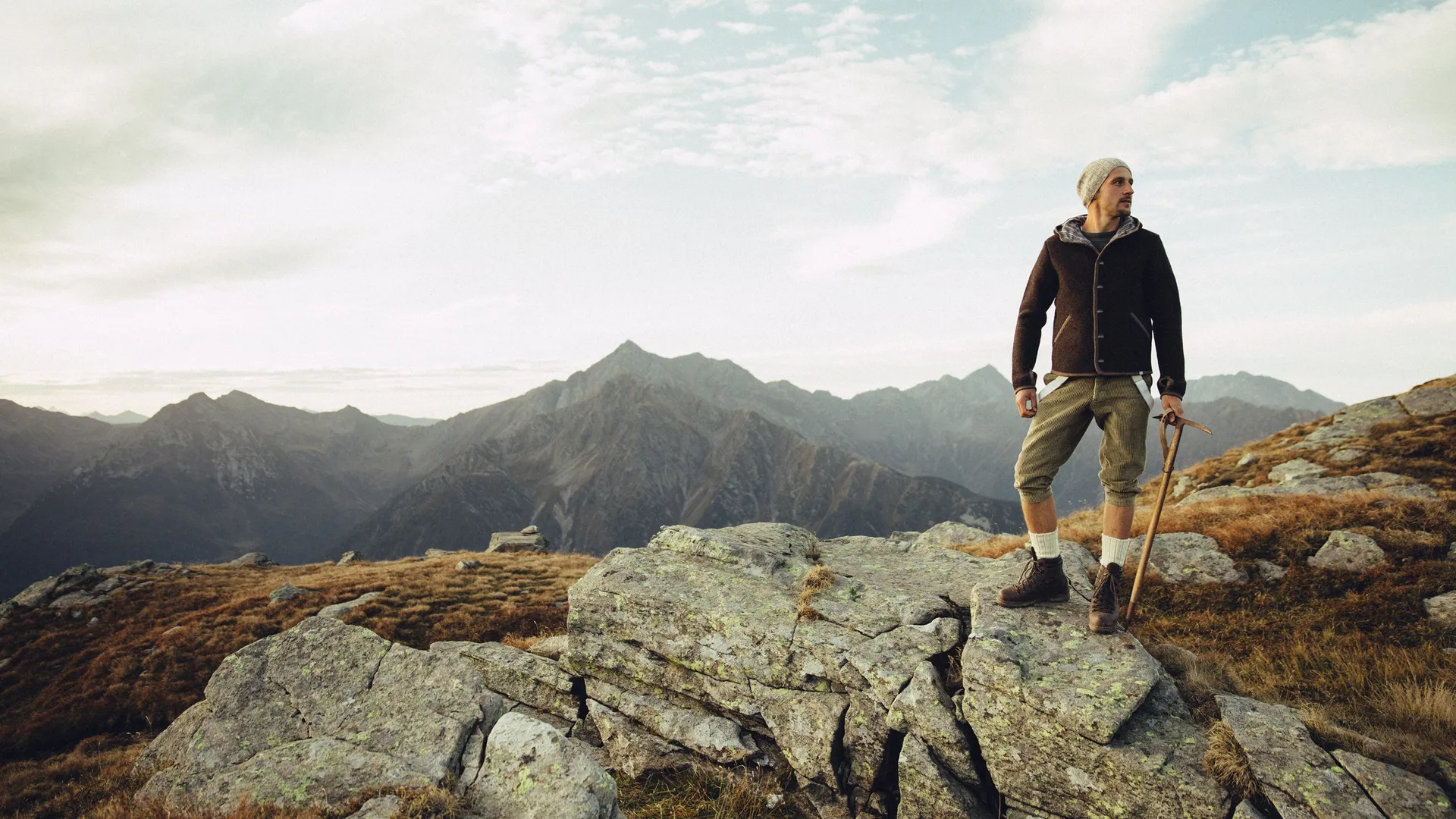 Wanderer in brauner Walkjacke und Kniebundhose mit Stock auf Felsen im Bergpanorama