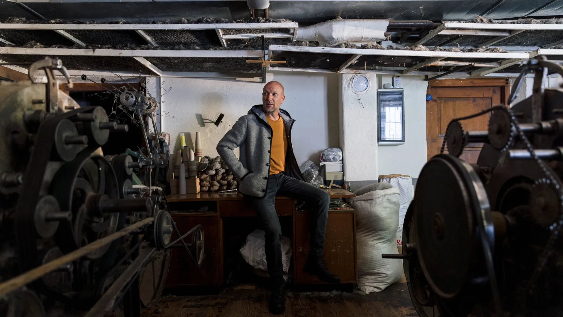 Mann in grauer Wolljacke und orangem Shirt sitzt zwischen alten Spinnmaschinen in der Werkstatt