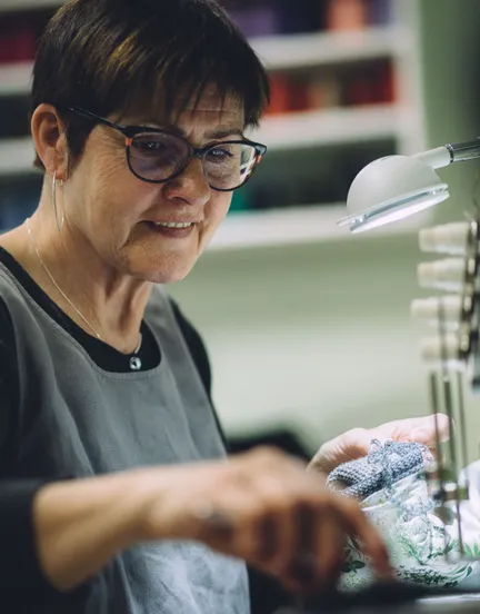 Handwerkerin mit Brille prüft unter Arbeitsleuchte konzentriert ein graues Strickteil in der Näherei