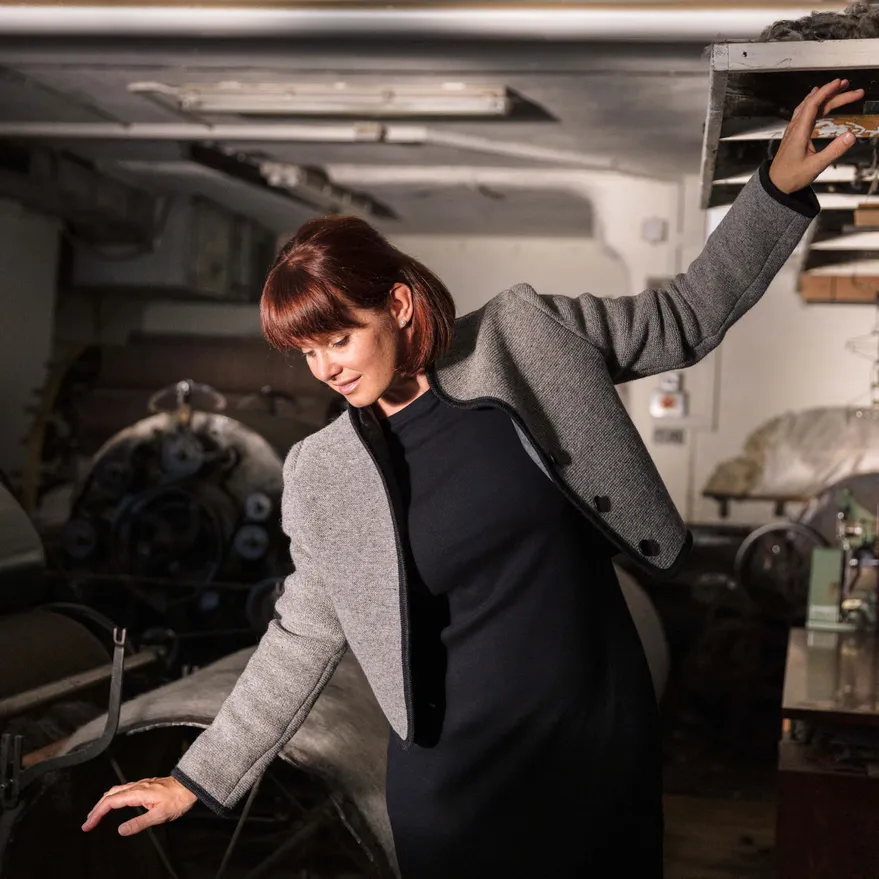 Frau in grauer Wolljacke und dunklem Kleid steht zwischen historischen Maschinen der Wollspinnerei