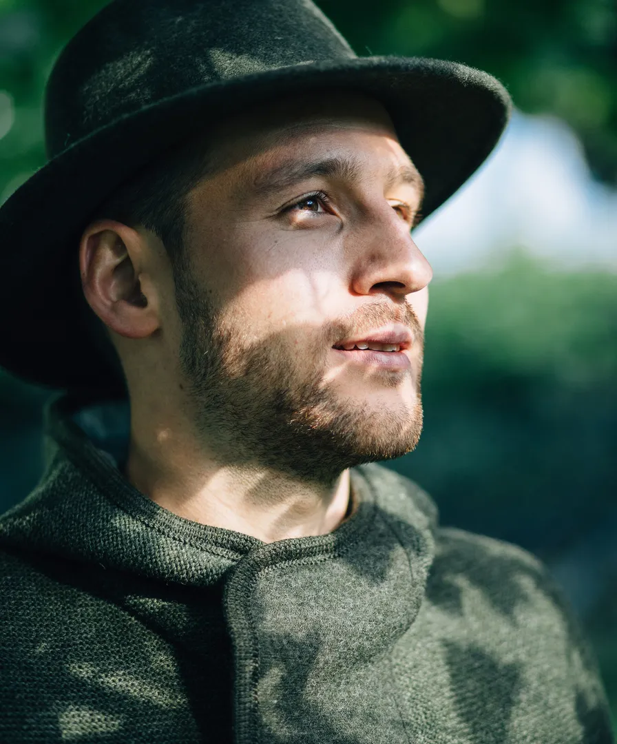 Mann mit dunkelgrünem Wollhut und Strickkapuzenjacke blickt in sonnige Ferne