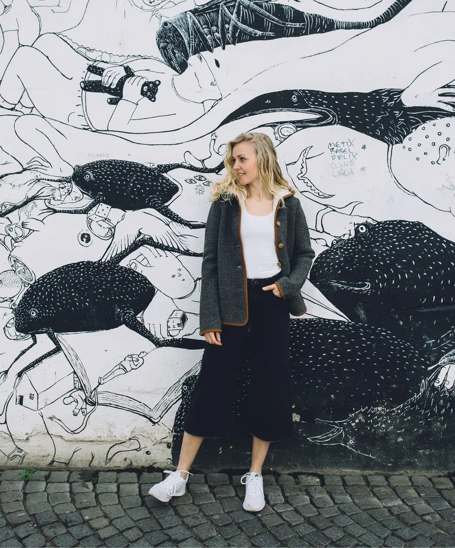 Blonde Frau in grauer Walkjanker mit brauner Paspelierung vor schwarz-weißem Graffiti-Mural