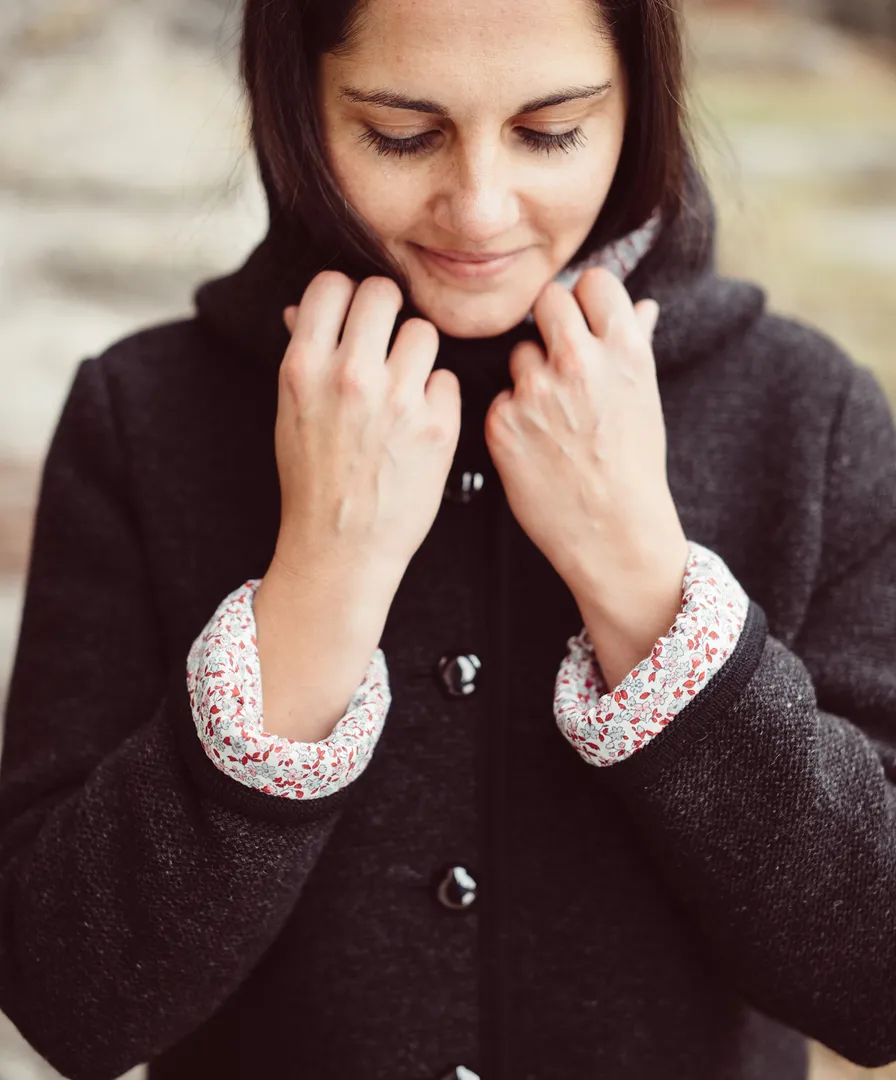 Dunkelhaarige Frau in dunkelgrauer Kapuzen-Walkjacke mit floraler Bluse, lächelnd