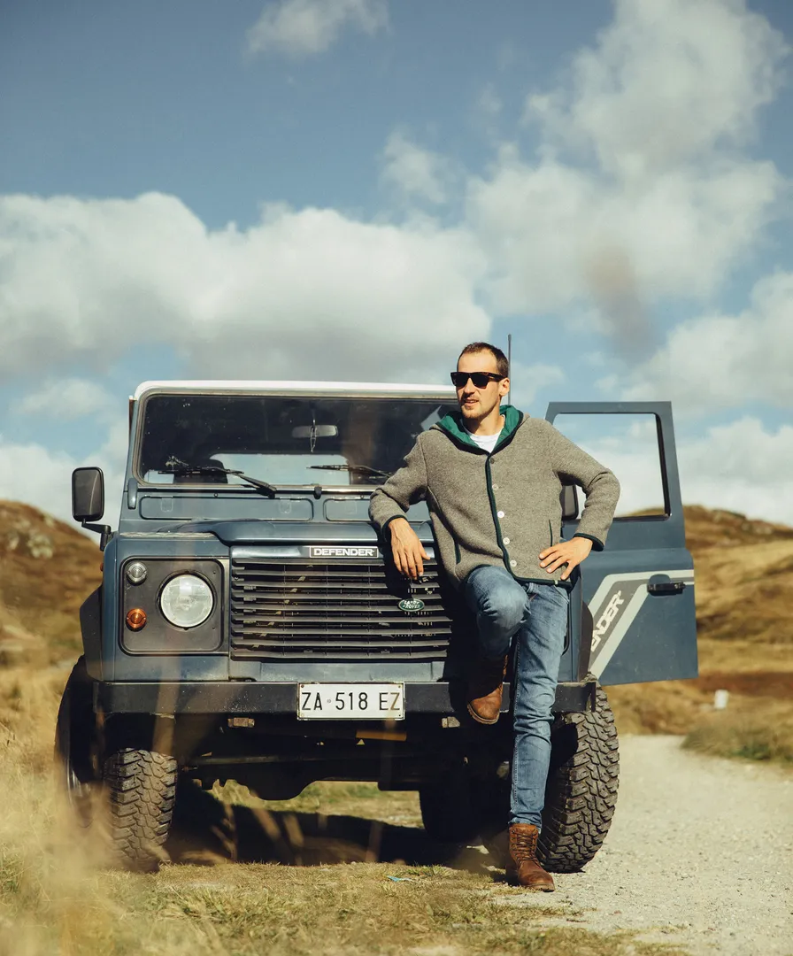 Mann in grauer Walkjacke mit grüner Kapuze lehnt an blauem Land Rover Defender in den Bergen