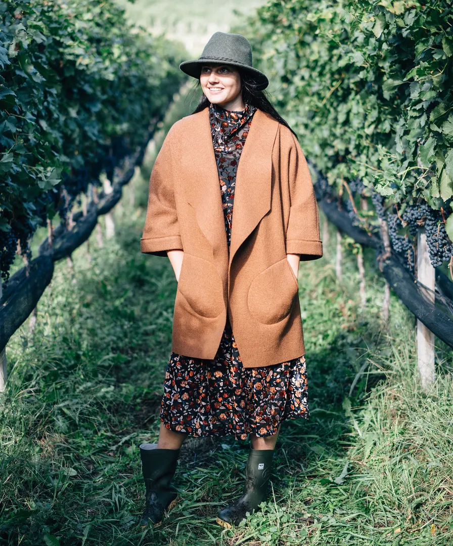 Frau in camelfarbenem Wollmantel, Blumenkleid und grünem Filzhut zwischen Weinreben im Weinberg